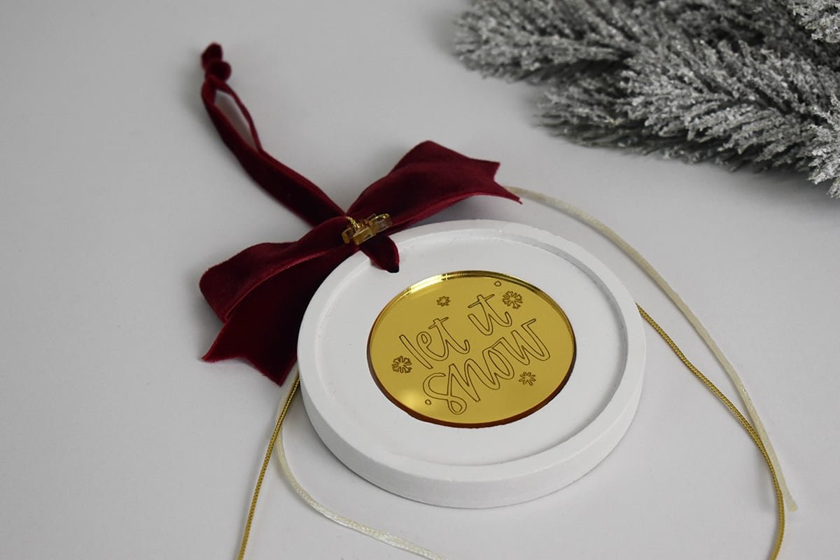 Let it Snow — Λευκό κυκλικό στολίδι με χρυσό σχέδιο και μπορντό κορδέλα.